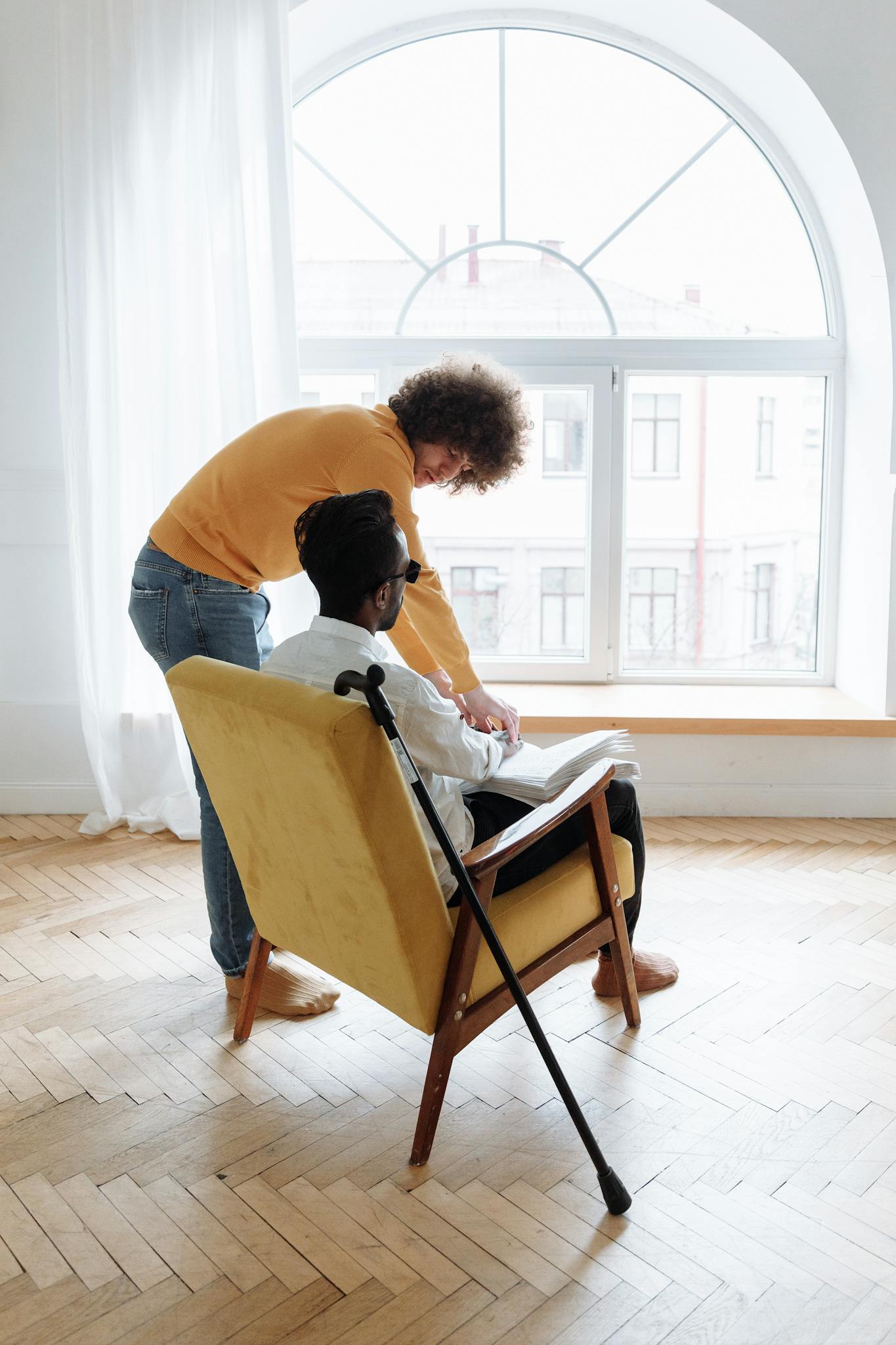 A mentor guides a visually impaired man in reading Braille indoors near a large window.