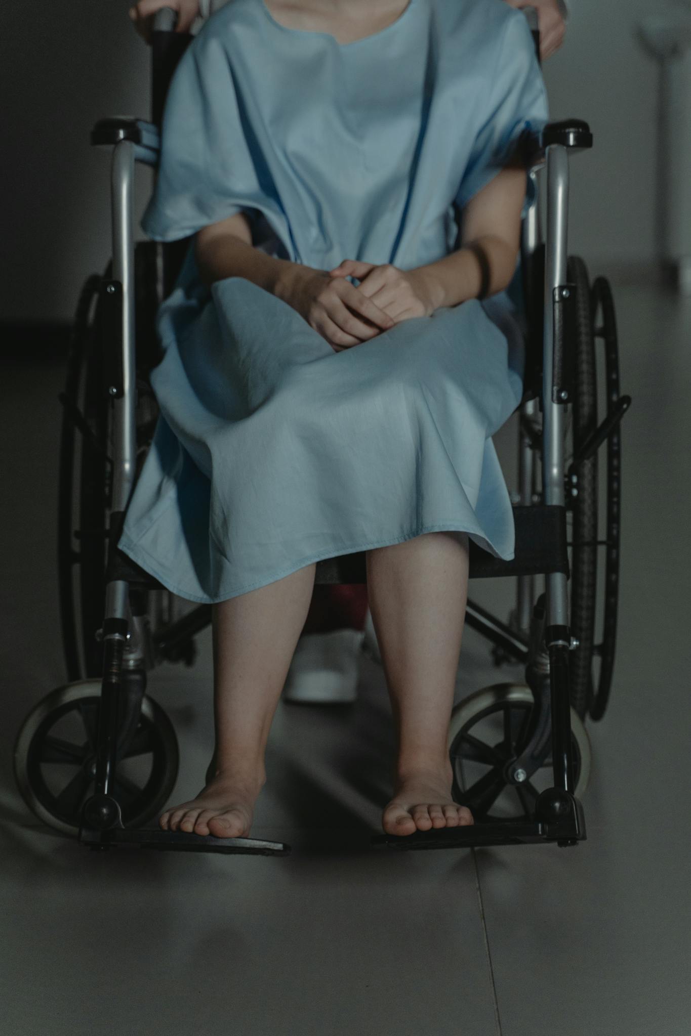 A young adult wearing a hospital gown sitting barefoot in a wheelchair indoors.