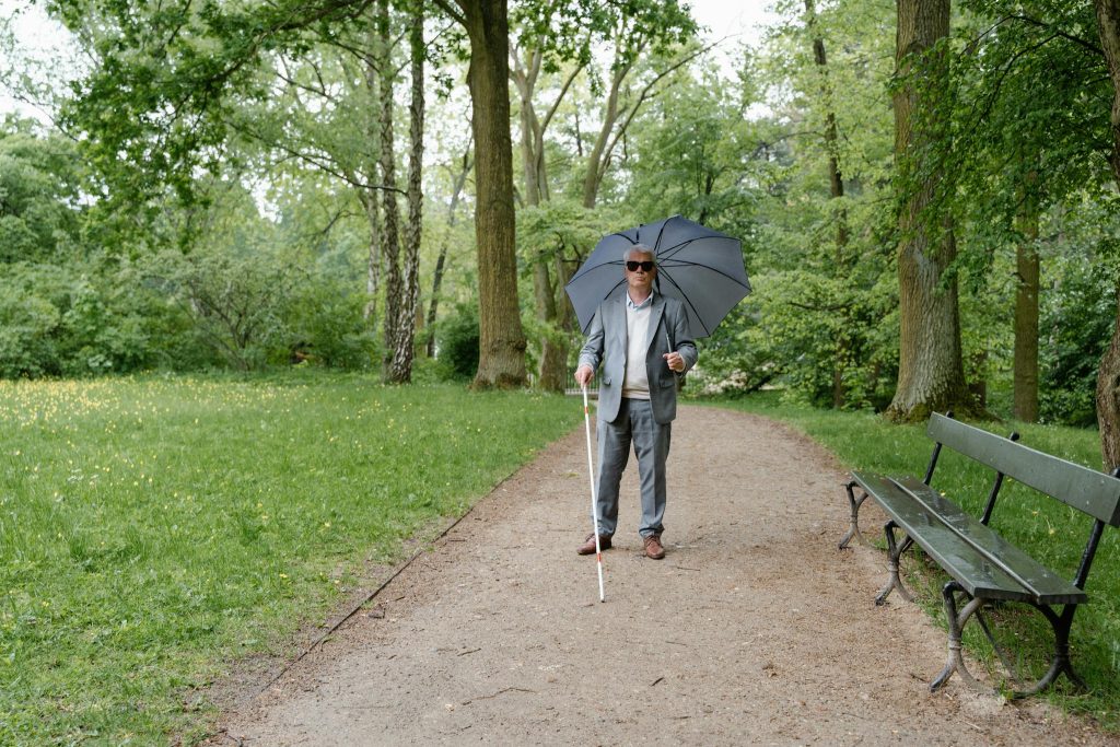 Elderly man with visual impairment walking with a cane and umbrella on a park pathway.
